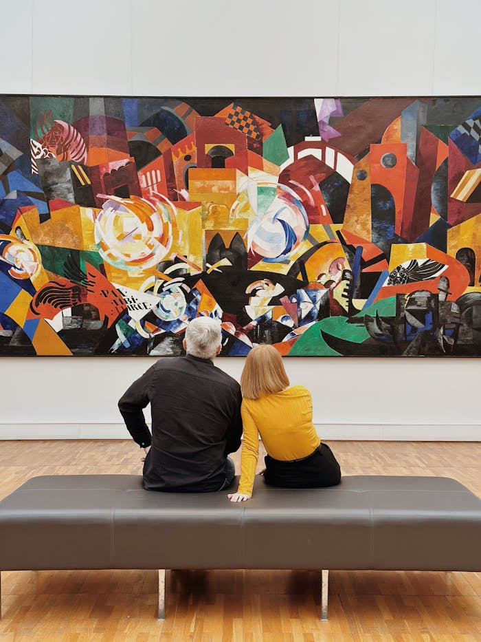 about-us A couple sits in a modern gallery, captivated by an abstract painting full of vibrant colors.